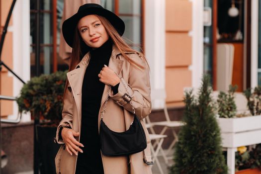 Stylish young woman in a beige coat and black hat and with a black purse on a city street. Women's street fashion. Autumn clothing.Urban style.