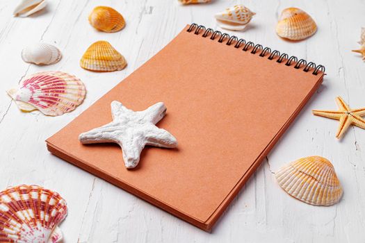 Brown open notepad surrounded by sea shells on white wooden table, copy space
