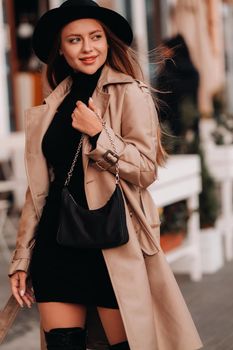 Stylish young woman in a beige coat and black hat and with a black purse on a city street. Women's street fashion. Autumn clothing.Urban style.