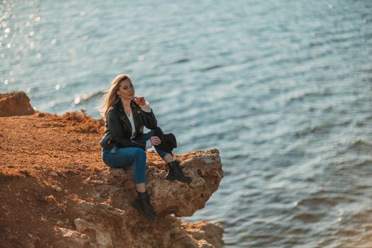 A blonde girl in a stylish black leather jacket is sitting on the seashore