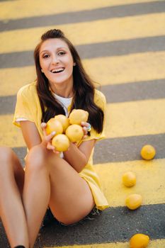 a girl with lemons in a yellow shirt, shorts and black shoes sits on a yellow pedestrian crossing in the city. The lemon mood.