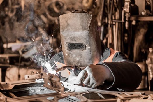A welder worker in a protective metal mask is engaged in welding and repair of a car door, metal work in the workshop of the plant.