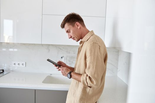 Hadsome smiling man is chilling on the minimalistic kitchen with smartphone