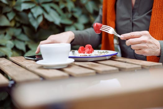 Mature woman eats delicious toast with cream and strawberries and takes mobile phone near cup at table on outdoors cafe terrace closeup