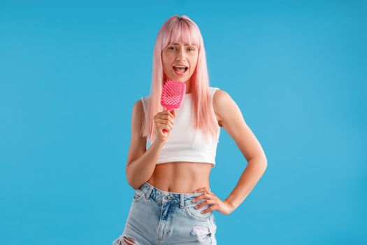 Joyful woman with pink hair holding hair comb and singing in it while standing isolated over blue studio background. Beauty, hair care, lifestyle concept