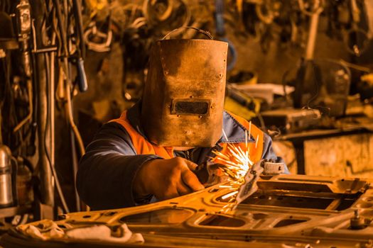 Welder worker in a protective iron mask repairs the car door and is engaged in welding work in the shop of the industrial plant.