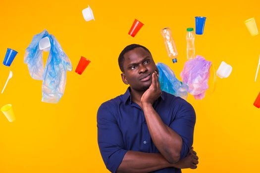 Problem of trash, plastic recycling, pollution and environmental concept - Serious african american man looking on trash on yellow background. He is thinking about ecology.