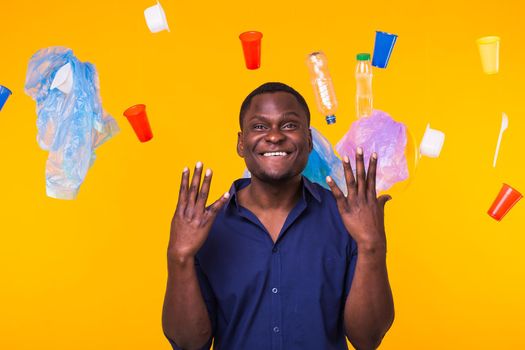 Problem of trash, plastic recycling, pollution and environmental concept - african american man is standing under trash on yellow background.