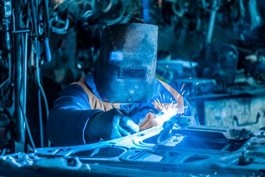 A male construction worker welder in a protective shield is engaged in welding work on metal and repairing a car door in the plant shop.