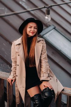 A stylish young woman in a beige coat and black hat sits on a rooftop in the city center. Women's street fashion. Autumn clothing.Urban style.