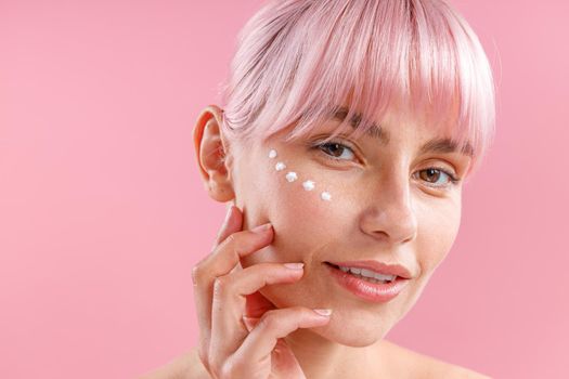 Portrait of charming woman with pink hair and face cream applied on her skin like dots isolated over pink background. Beauty, spa, skin care concept