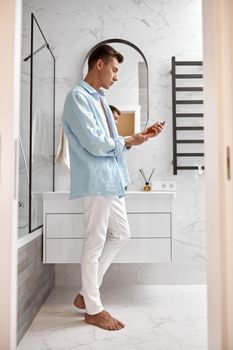 Photo of adult caucasian beautiful man in front of mirror in light modern bathroom