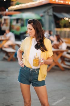 a girl in a yellow shirt and shorts with a glass of lemon juice is standing on the street.