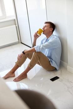 Caucasian hadsome young man in blue shirt is sitting on the floor and chilling at modern kitchen