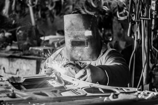 The welder in the protective shield is engaged in welding work on iron, and repair of the automobile door to the workshop of the industrial plant.