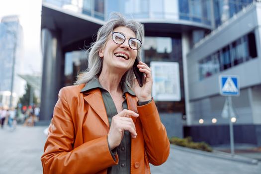 Joyful hoary haired senior lady with spectacles in leather jacket talks by cellphone on modern city street on autumn day