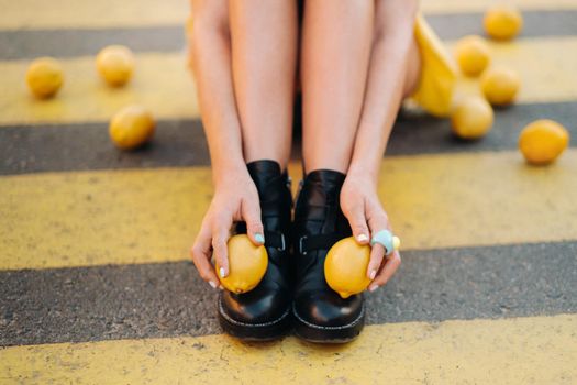 a girl with lemons in a yellow shirt, shorts and black shoes sits on a yellow pedestrian crossing in the city. The lemon mood.