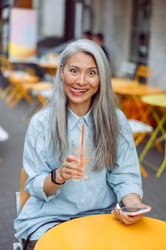 Mature Asian woman with glass of water and smartphone makes funny face sitting at small table on outdoors cafe terrace