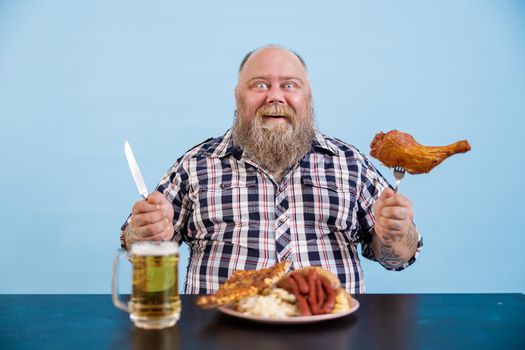 Smiling mature man with overweight holds fork with chicken leg sitting at table with greasy food and beer on light blue background in studio