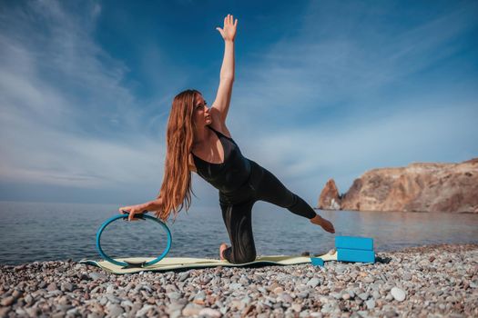 Young woman with long hair, fitness instructor in black Sportswear Leggings and Tops, stretching on a yoga mat with magic pilates ring near the sea on a sunny day, female fitness yoga routine concept.