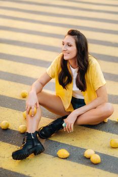 a girl with lemons in a yellow shirt, shorts and black shoes sits on a yellow pedestrian crossing in the city. The lemon mood.