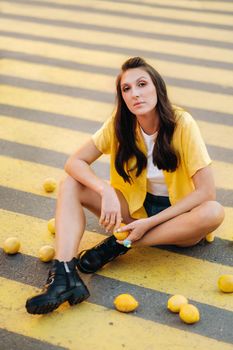 a girl with lemons in a yellow shirt, shorts and black shoes sits on a yellow pedestrian crossing in the city. The lemon mood.