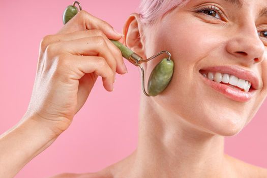 Close up portrait of lovely woman with fresh skin smiling and using natural jade facial roller for skin care isolated over pink background. Face massage and beauty trends concept