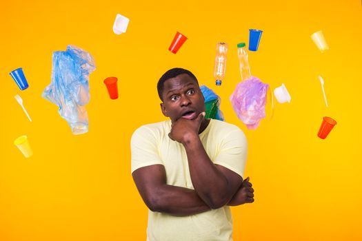 Problem of trash, plastic recycling, pollution and environmental concept - Serious african american man looking on trash on yellow background. He is thinking about ecology.