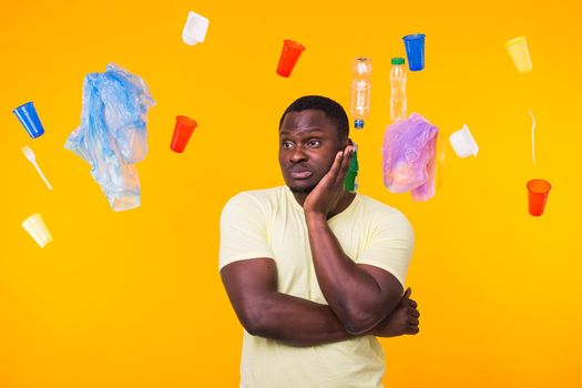 Problem of trash, plastic recycling, pollution and environmental concept - Serious african american man looking on trash on yellow background. He is thinking about ecology.