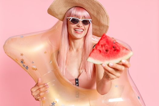 Excited woman with pink hair in swimsuit and sunglasses holding watermelon slice and inflatable star for swimming isolated over pink background. Summer vacation, fun, party concept