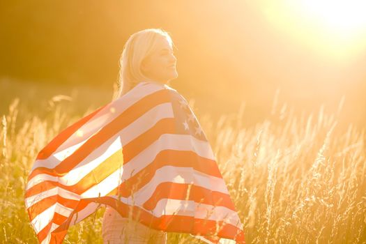 Beautiful young girl holding an American flag in the wind in a field of rye. Summer landscape against the blue sky. Horizontal orientation.