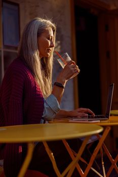 Senior Asian woman with silver hair holds glass sitting in front of laptop at small round table on outdoors cafe terrace at sunset side view