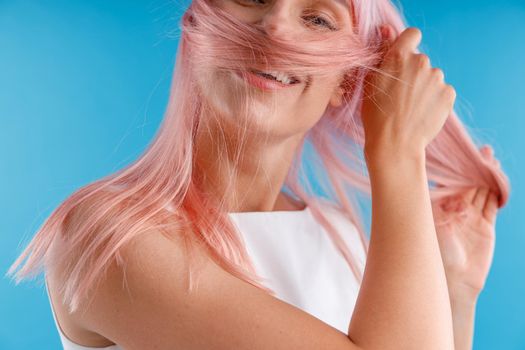 Portrait of beautiful female model playing with pink hair, wrapping it around her face while posing isolated over blue studio background. Beauty, hair care concept