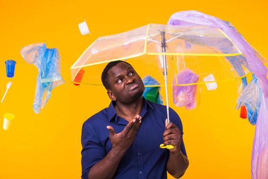 Problem of trash, plastic recycling, pollution and environmental concept - Serious african american man looking on trash on yellow background. He is thinking about ecology.