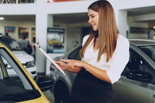 Attractive young female car dealer standing in showroom near a new car
