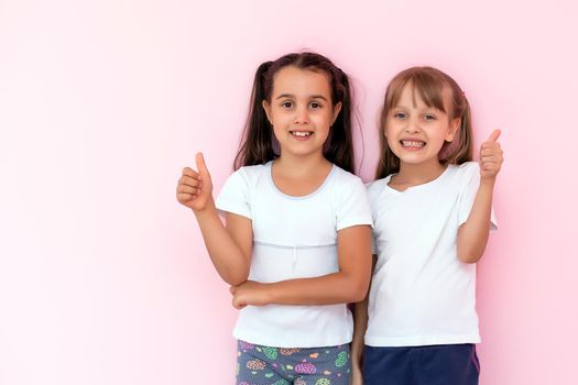 Children showing like. Two adorable happy little girls gesturing thumbs up together and smiling to camera, excellent feedback, good job. indoor studio shot isolated on pink background