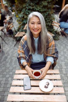 Positive long haired mature Asian woman with cup of fresh tea, candies and mobile phone sits at table on outdoors cafe terrace on autumn day