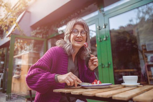 Smiling grey haired senior woman guest in purple jacket eats tasty strawberry dessert sitting at table on outdoors cafe terrace on autumn day