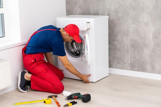 Plumber in overalls with tools is repairing a washing machine in the house