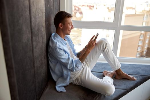 Happy adult caucasian male is sitting near panoramic window and chilling