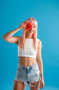 Smiling young woman with pink hair holding a slice of watermelon over her eye, standing isolated over blue studio background. Beauty and health concept