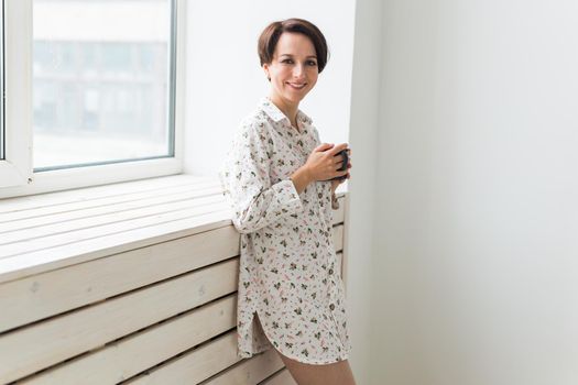 Beautiful young woman with cup of tea standing near the window at home