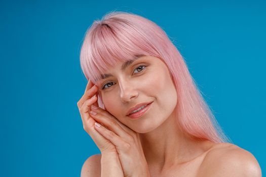 Beautiful naked woman with pink hair and perfect skin smiling at camera, leaning her head on her hands, posing isolated over blue studio background. Beauty, skin care concept