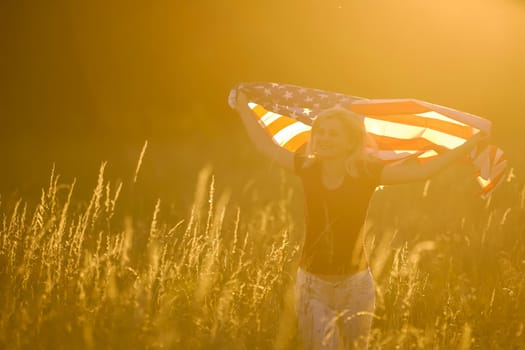 Beautiful young girl holding an American flag in the wind in a field of rye. Summer landscape against the blue sky. Horizontal orientation.