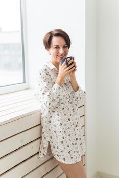 Beautiful young woman with cup of tea standing near the window at home