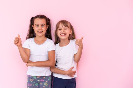 Children showing like. Two adorable happy little girls gesturing thumbs up together and smiling to camera, excellent feedback, good job. indoor studio shot isolated on pink background