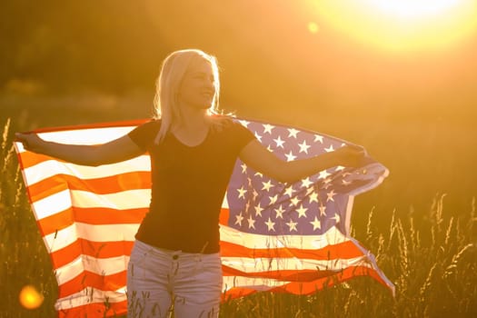 Beautiful young girl holding an American flag in the wind in a field of rye. Summer landscape against the blue sky. Horizontal orientation.