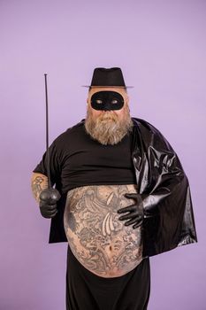 Funny middle aged gentleman with overweight in Zorro costume shows large bare tummy with different tattoos standing on purple background in studio