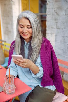 Pretty senior Asian lady with grey hair reads sms on mobile phone holding drink at table on outdoors cafe terrace