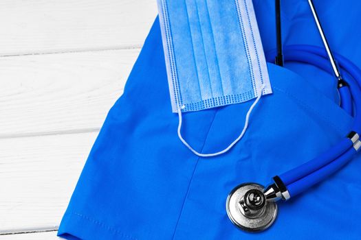 Doctor medical uniform with stethoscope and face mask on white wooden background close up